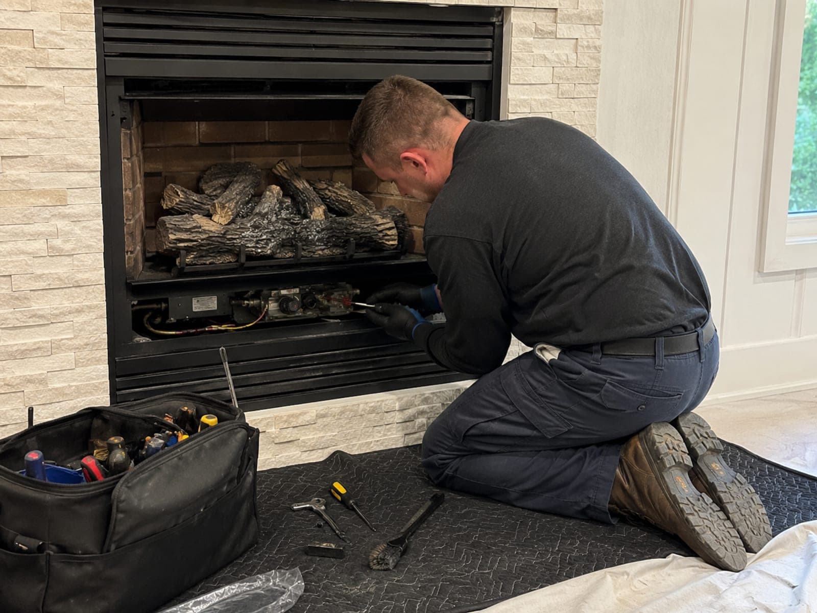 Chimney technician kneeling on a drop mat servicing the valve train under a factory-built gas fireplace, with the ceramic log set and burner visible above